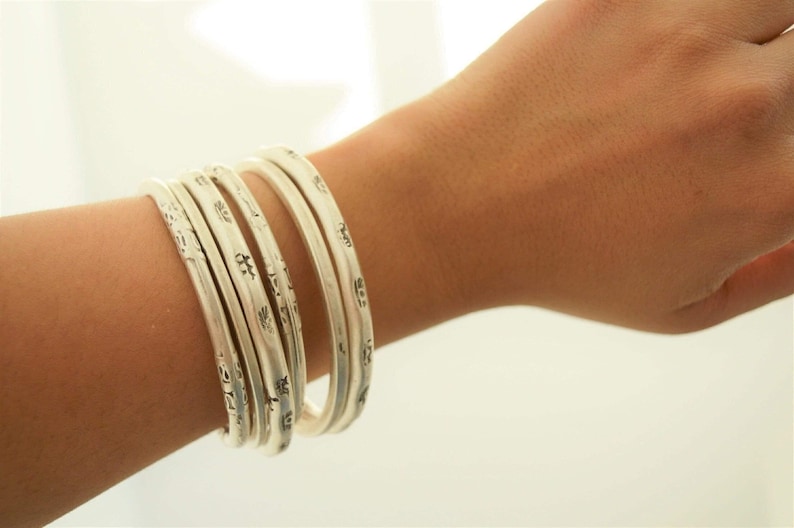 May include: A set of six silver bangles with etched designs. The bracelets are stacked on a wrist, showcasing their intricate patterns and reflective surface. The jewelry is displayed against a soft, neutral background.