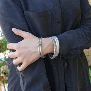 May include: Two silver bracelets are worn on a person's wrist. One bracelet has a decorative pattern, and the other has a series of decorative elements. The person is wearing a dark gray button-down shirt.