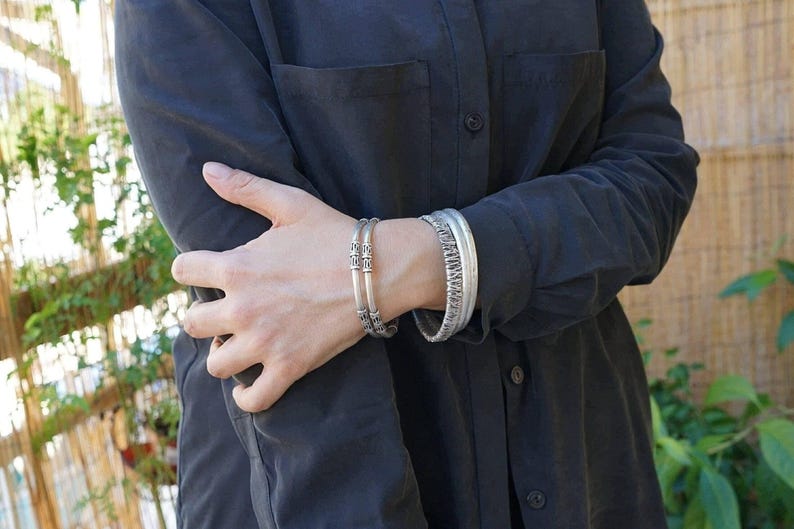May include: Two silver bracelets are worn on a person's wrist. One bracelet has a decorative pattern, and the other has a series of decorative elements. The person is wearing a dark gray button-down shirt.