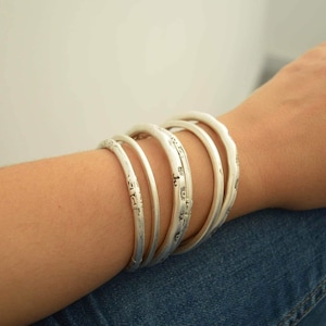May include: A set of six silver-colored bangles with a textured, slightly irregular surface. The bracelets are stacked on a person's arm, with the arm resting on blue denim jeans. The bangles have a simple, elegant design.