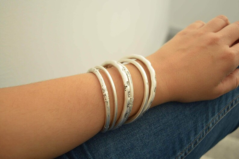 May include: A set of six silver-colored bangles with a textured, slightly irregular surface. The bracelets are stacked on a person's arm, with the arm resting on blue denim jeans. The bangles have a simple, elegant design.