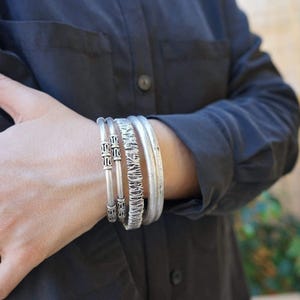 May include: A set of silver bracelets with various designs. The bracelets include a textured band, a band with geometric patterns, and two simple bands. The bracelets are worn on the wrist, against a dark gray shirt.
