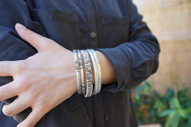 May include: A set of silver bracelets with various designs. The bracelets include a textured band, a band with geometric patterns, and two simple bands. The bracelets are worn on the wrist, against a dark gray shirt.