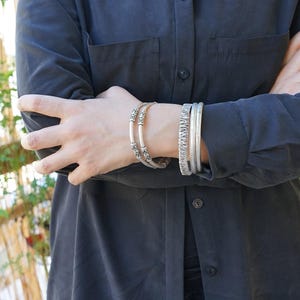 May include: Silver bracelets adorn an arm, showcasing a variety of designs. One bracelet features intricate patterns, while others have a textured appearance. The bracelets are worn against a dark shirt, creating a contrast that highlights the jewelry.