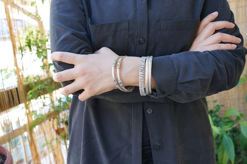 May include: Silver bracelets adorn an arm, showcasing a variety of designs. One bracelet features intricate patterns, while others have a textured appearance. The bracelets are worn against a dark shirt, creating a contrast that highlights the jewelry.