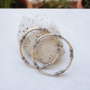 May include: Two silver bangles with decorative rectangular accents. The bangles are resting on a light-colored, textured stone. The background is a white surface with hints of greenery.
