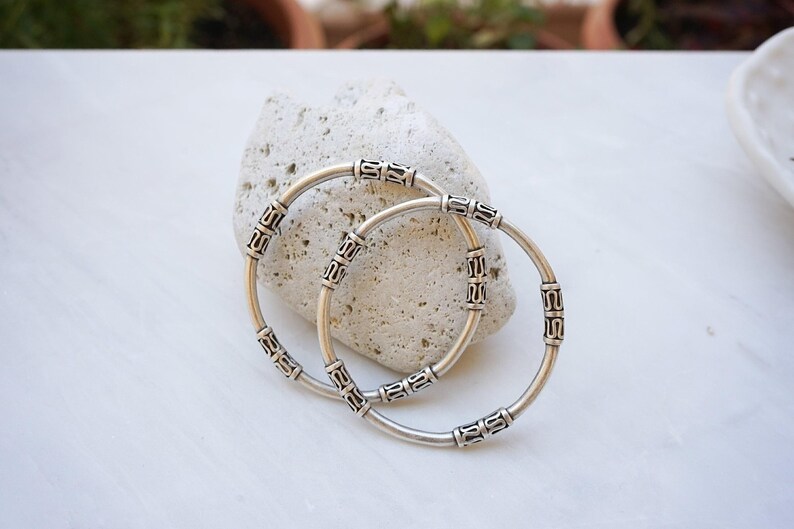 May include: Two silver bangles with decorative rectangular accents. The bangles are resting on a light-colored, textured stone. The background is a white surface with hints of greenery.