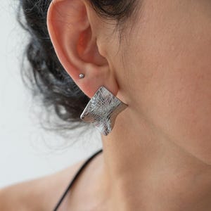 Puede incluir: Primer plano de un pendiente cuadrado plateado y texturizado. El pendiente se lleva en la oreja de una persona con cabello oscuro. El pendiente tiene un diseño abstracto único.