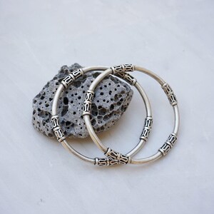 May include: Two silver-colored bangles with decorative patterns rest on a porous gray stone. The bracelets are circular, with detailed designs around their circumference. The background is a light, marbled surface, providing a neutral backdrop for the jewelry.