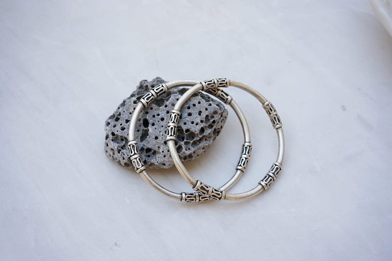 May include: Two silver-colored bangles with decorative patterns rest on a porous gray stone. The bracelets are circular, with detailed designs around their circumference. The background is a light, marbled surface, providing a neutral backdrop for the jewelry.