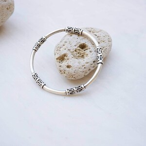May include: A silver bracelet with decorative rectangular accents. The bracelet is circular and rests on a white surface next to a light-colored stone. The bracelet's design features repeating patterns.