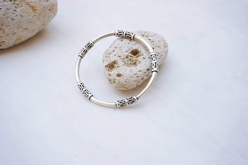 May include: A silver bracelet with decorative rectangular accents. The bracelet is circular and rests on a white surface next to a light-colored stone. The bracelet's design features repeating patterns.