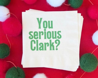 May include: A pile of cream-coloured napkins printed with the green text "You serious Clark?". The napkins are set against a red backdrop and surrounded by a garland of red, white, and green felt balls.
