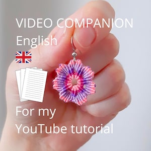 Puede incluir: Un pendiente de macramé con un diseño circular en forma de flor, hecho con hilo multicolor en tonos rosa, morado y blanco. El pendiente cuelga de un gancho de metal plateado. Texto en la imagen: VIDEO COMPANION English For my YouTube tutorial.