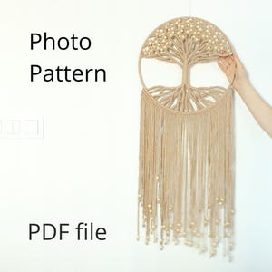 Op de afbeelding: Een beige macramé wandhanger in de vorm van een levensboom met houten kralen. De wandhanger heeft een franje van kwastjes en hangt aan een witte muur. De tekst "Photo Pattern PDF file" staat onder de wandhanger.