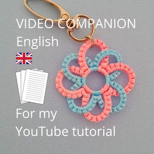 May include: A keychain featuring a flower-shaped macrame charm made of alternating light blue and coral pink cord, attached to a gold-toned metal lobster clasp. The image includes the text: VIDEO COMPANION English, a flag icon, paper icons, and For my YouTube tutorial.