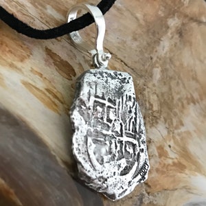 Puede incluir: Collar con colgante de plata que representa una r&eacute;plica de una moneda espa&ntilde;ola de ocho reales. La moneda tiene un dise&ntilde;o en relieve y est&aacute; ligeramente desgastada. El colgante est&aacute; unido a un cord&oacute;n negro.