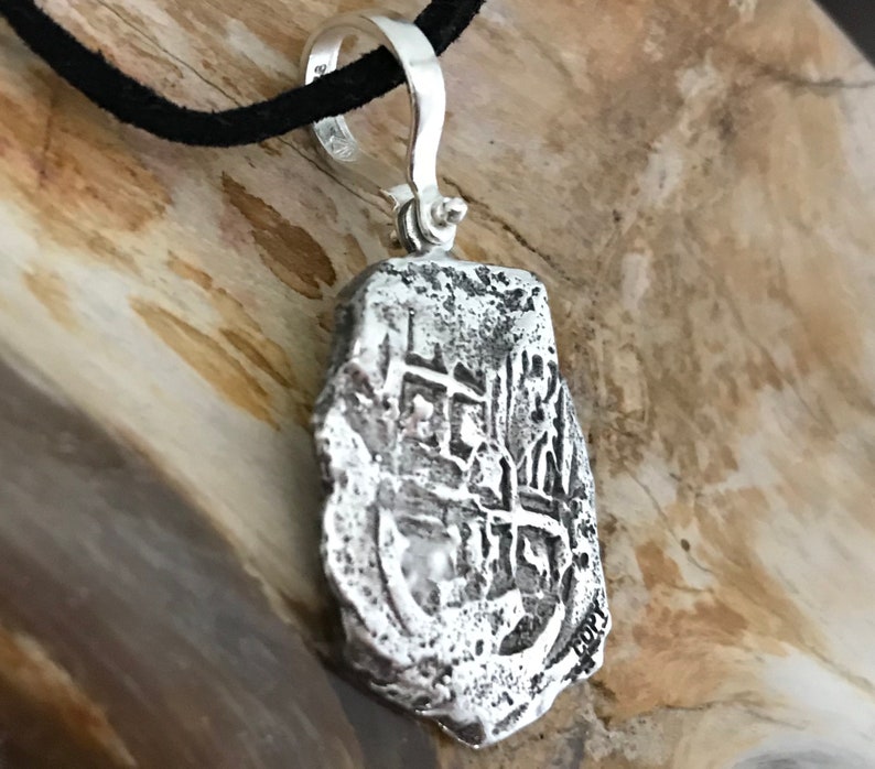 Puede incluir: Collar con colgante de plata que representa una r&eacute;plica de una moneda espa&ntilde;ola de ocho reales. La moneda tiene un dise&ntilde;o en relieve y est&aacute; ligeramente desgastada. El colgante est&aacute; unido a un cord&oacute;n negro.