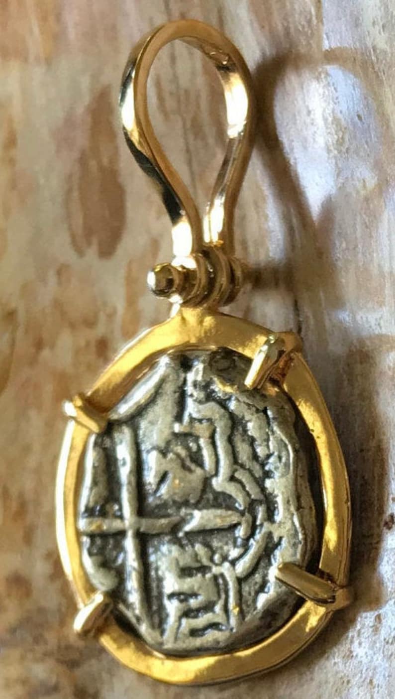May include: A gold pendant featuring a silver coin with a cross and other markings. The coin is set in a gold frame with a bail loop.