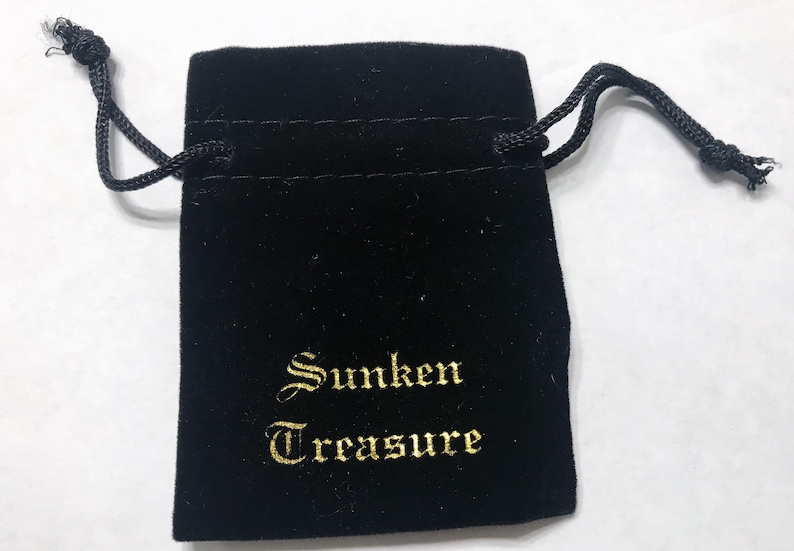 Puede incluir: Una bolsa de terciopelo negro con cord&oacute;n de ajuste y bordado dorado que dice "Sunken Treasure".