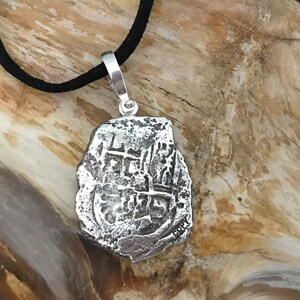 Puede incluir: Collar con colgante de plata que representa una r&eacute;plica de una moneda espa&ntilde;ola de ocho reales. La moneda tiene un dise&ntilde;o de cruz y la inscripci&oacute;n "8 R", y est&aacute; en un cord&oacute;n negro.