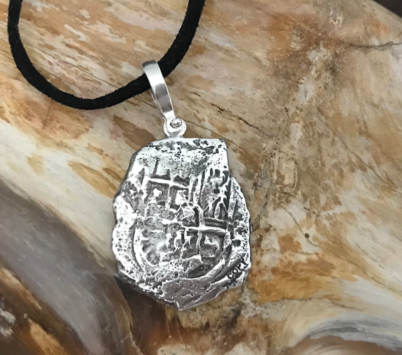 Puede incluir: Collar con colgante de plata que representa una r&eacute;plica de una moneda espa&ntilde;ola de ocho reales. La moneda tiene un dise&ntilde;o de cruz y la inscripci&oacute;n "8 R", y est&aacute; en un cord&oacute;n negro.