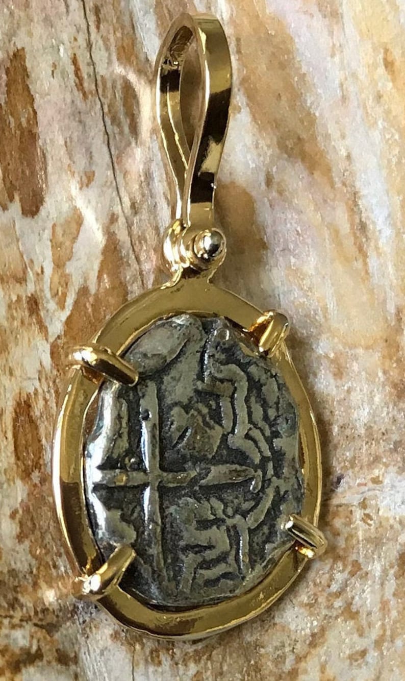 May include: A silver coin pendant with a gold frame and bail. The coin features a cross and other markings.