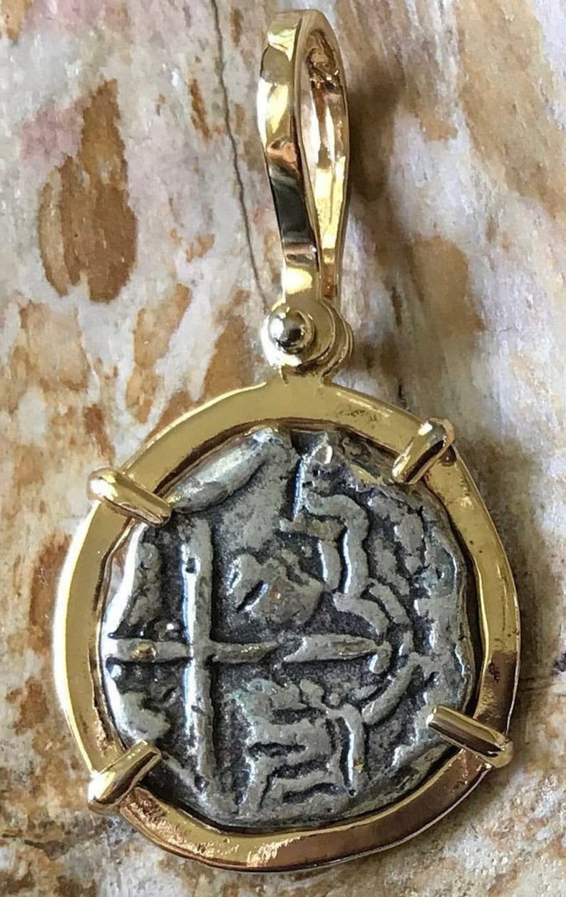 May include: A silver coin pendant with a gold frame. The coin features a cross and a design of a person on horseback. The pendant is on a wooden surface.