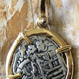 May include: A silver coin pendant with a gold frame and a gold bail. The coin features a design of a person's head and shoulders with text around the edge. The pendant is approximately 2.5 cm in diameter.