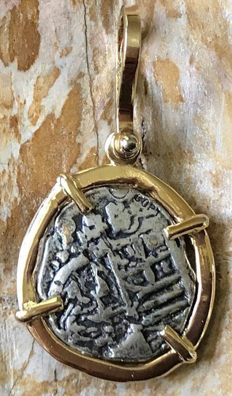May include: A silver coin pendant with a gold frame and a gold bail. The coin features a design of a person's head and shoulders with text around the edge. The pendant is approximately 2.5 cm in diameter.