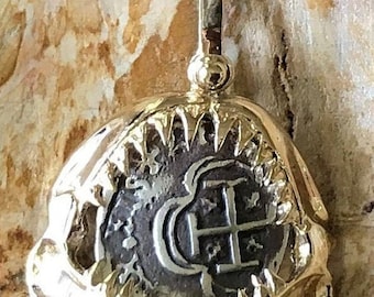 Op de afbeelding: Een gouden hanger met een zilveren munt met een kruis en de letter 'C' in een cirkel. De munt is ingezet in een gouden frame met een gekartelde rand.