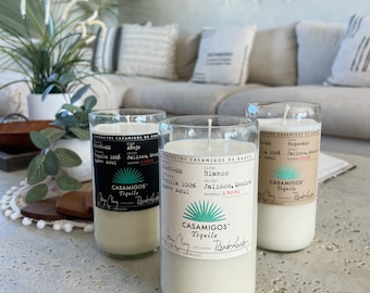 May include: Three clear glass candle jars with white wax and black labels. The labels read "Casamigos Tequila" and include details about the tequila, such as the class, origin, and proof. The candles are sitting on a light grey textured surface.