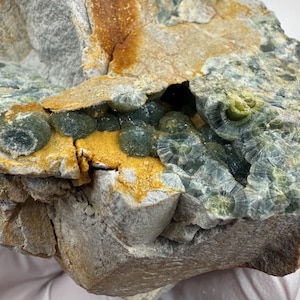 May include: A close-up of a rock formation with a variety of colors and textures. The rock has a gray base with patches of orange, yellow, and green. The green areas have a crystalline appearance, and the rock is held by a gloved hand.