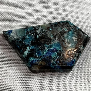 May include: A dark blue and green gemstone with a unique, irregular shape. The stone has a mottled appearance with swirls of color.