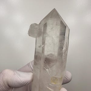 May include: A clear quartz crystal cluster with a pointed top and several smaller crystal formations attached. The crystal is held by a gloved hand, showcasing its natural clarity and unique structure. The crystal is a natural mineral specimen.