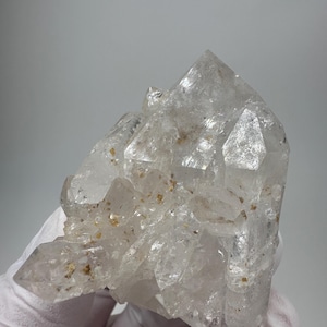 Pode incluir: Um aglomerado de cristais de quartzo transparente com múltiplas pontas e uma superfície áspera e texturizada. O cristal tem inclusões de pequenas partículas douradas. O quartzo é segurado por uma mão enluvada contra um fundo neutro.