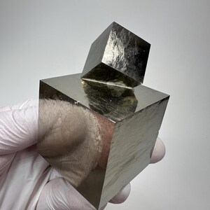 以下が含まれることがあります： 2つの金属製の黄鉄鉱の立方体で、1つがもう1つの上に乗っています。立方体は反射性のある銀色の光沢があり、白い背景に対して手袋をはめた手で保持されています。立方体の表面には自然なテクスチャが見られます。