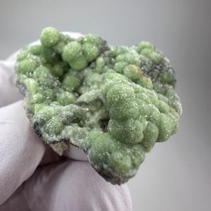 May include: A close-up of a light green mineral specimen. The mineral has a bubbly, textured surface with a dark gray base. The specimen is held by a gloved hand against a plain background.