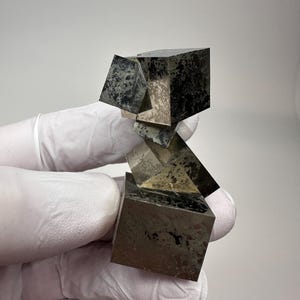 Może przedstawiać: Skupisko metalicznych kryształów pirytu o wyraźnych sześciennych formacjach, z odblaskowymi, mosiężnymi powierzchniami oraz ciemnymi, teksturowanymi wtrąceniami mineralnymi. Okaz wykazuje ostre geometryczne krawędzie i ułożoną strukturę.