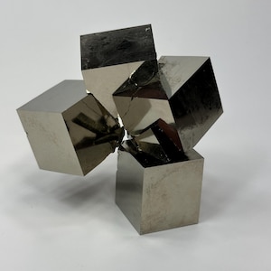May include: A metallic sculpture featuring four interconnected, cube-shaped forms. The cubes have a reflective, silver-grey surface with visible facets and slight imperfections. The arrangement suggests a modern, abstract design.