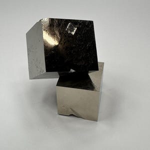 Może przedstawiać: Dwa metaliczne, srebrzyste, sześcienne okazy minerałów ułożone jeden na drugim. Górny sześcian jest ustawiony pod kątem, ukazując gładką, odblaskową powierzchnię z małym, wyraźnym wgłębieniem w kształcie rombu na górnej ściance.