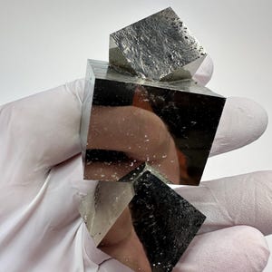 Może przedstawiać: Wyświetlane są trzy kostki pirytu, minerału znanego również jako złoto głupców. Kostki mają metaliczną, odblaskową powierzchnię i są ułożone jedna na drugiej. Minerał jest trzymany przez dłoń w rękawicy.