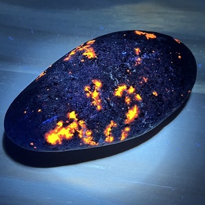 Puede incluir: Una roca de color gris oscuro, de forma ovalada, con motas brillantes de color naranja y amarillo. La roca está iluminada con luz UV, lo que hace que las motas brillen.