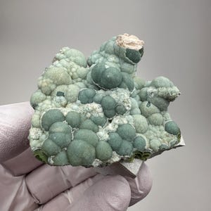 May include: A mineral specimen featuring a cluster of rounded, botryoidal formations in shades of sage green and forest green. The surface texture is bumpy and granular, with some areas showing a lighter, pale green crystalline coating.