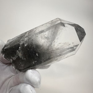 May include: A clear quartz crystal with black inclusions, also known as smoky quartz, in a double terminated point shape. The crystal is being held in a gloved hand.