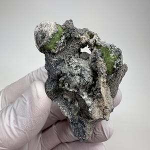 以下が含まれることがあります： 2つの鮮やかな緑色の結晶を含む、ざらざらとした質感の岩石。岩は主にグレーで、白とベージュのアクセントがあります。背景は無地の白です。