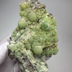 May include: A light green mineral specimen with a bubbly, rounded texture. The surface is covered in small, spherical formations, with larger, more prominent orbs. The base of the specimen is a darker, rougher rock.