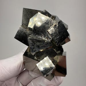 Może przedstawiać: Gromada kryształów pirytu, znanego również jako złoto głupców, o metalicznej, odblaskowej powierzchni. Sześcienne kryształy są zgrupowane, tworząc unikalny i przyciągający wzrok okaz minerału. Kolor jest błyszczący, srebrzysto-złoty.