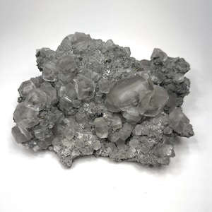 Pode incluir: Um aglomerado de cristais minerais cúbicos, translúcidos e incolores, incrustados numa matriz cinzenta, rugosa e texturada. O espécime apresenta numerosas formações geométricas de vários tamanhos, criando uma superfície irregular.