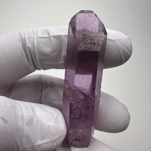 Puede incluir: Cristal de amatista facetado y alargado. El cristal es de color púrpura translúcido con algunas inclusiones internas. El cristal está sostenido por una mano enguantada sobre un fondo blanco. La forma del cristal es un prisma de seis lados.